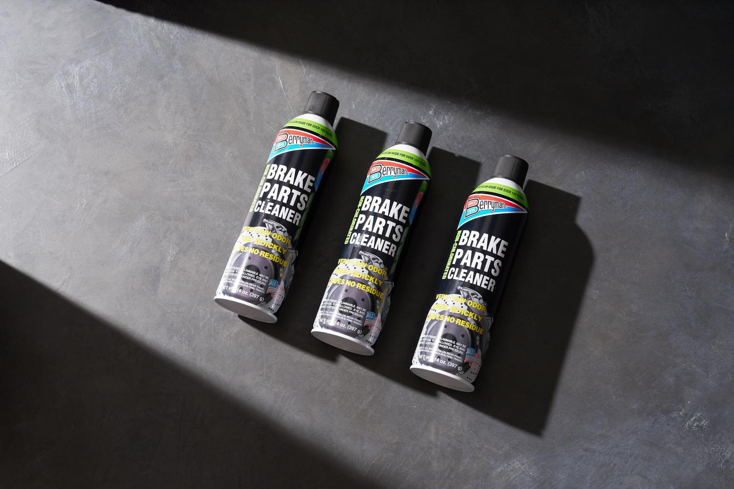 Three aerosol cans of Berryman Brake Parts Cleaner arranged in a diagonal line on a textured gray surface, shot from above with dramatic directional lighting casting strong shadows. The black cans feature yellow warning text and colorful branded l...