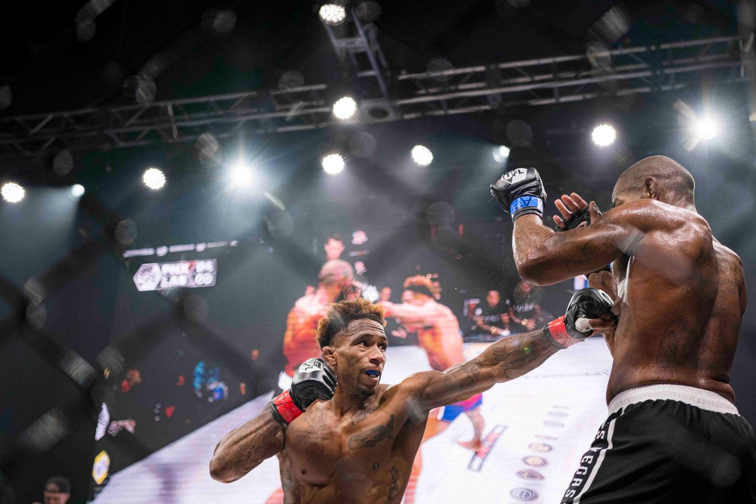 Two MMA fighters engaged in combat inside a professional fighting cage, with dramatic overhead stage lighting and visible crowd in the background. The fighter on the left is extending a punch while the opponent on the right is in defensive positio...