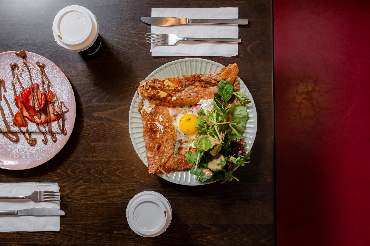 Overhead flat-lay composition of a restaurant table featuring a savory galette with egg and crispy fried chicken on a ridged ceramic plate, alongside a strawberry dessert crepe on a pink plate, with coffee cups and napkin-wrapped silverware arrang...