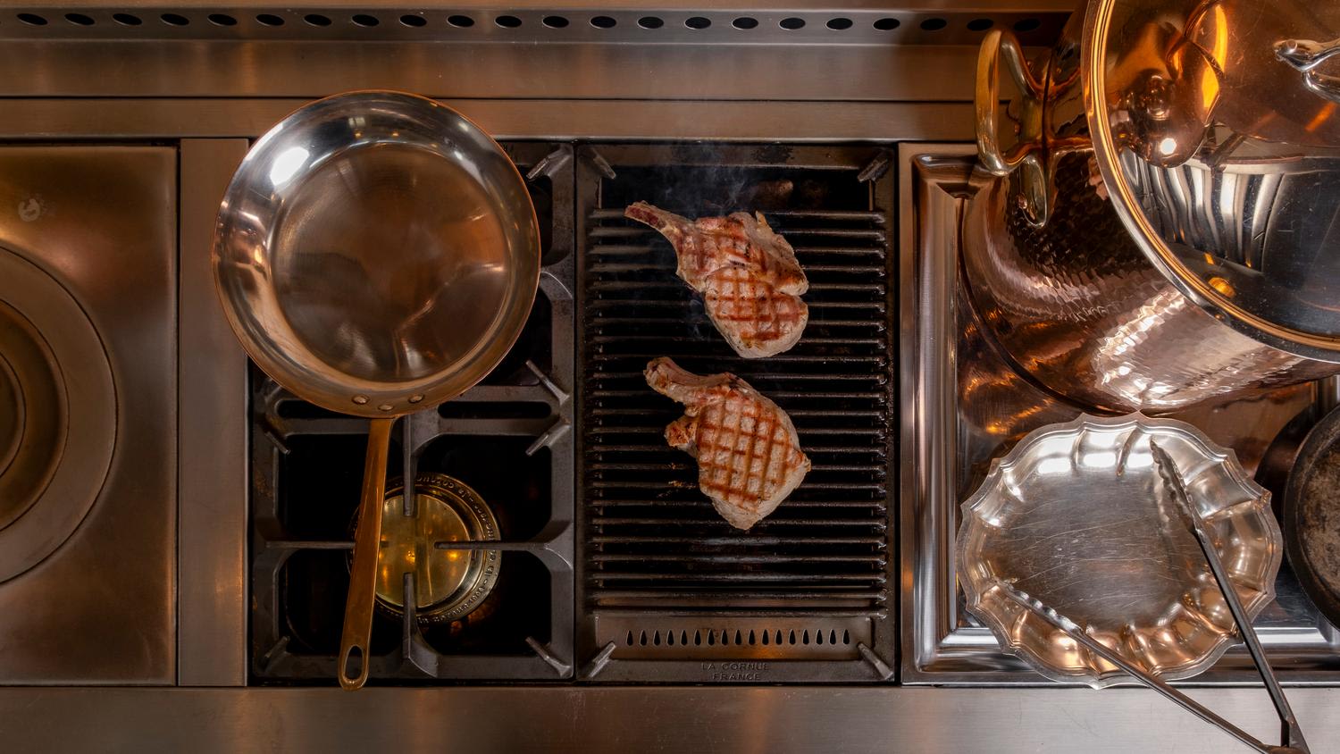 Overhead view of a professional kitchen range featuring two grilled pork chops with crosshatch marks on a built-in grill, with copper and stainless cookware arranged on either side, including a sauté pan, serving platters, and utensils on a stainl...