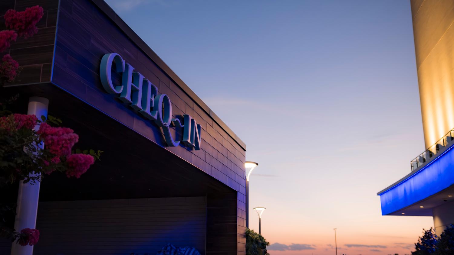 Modern entertainment venue exterior at dusk with illuminated 'CHECK IN' signage on building facade. The building features contemporary architecture with clean lines, blue accent lighting, pink flowering plants in foreground planters, and streetlig...