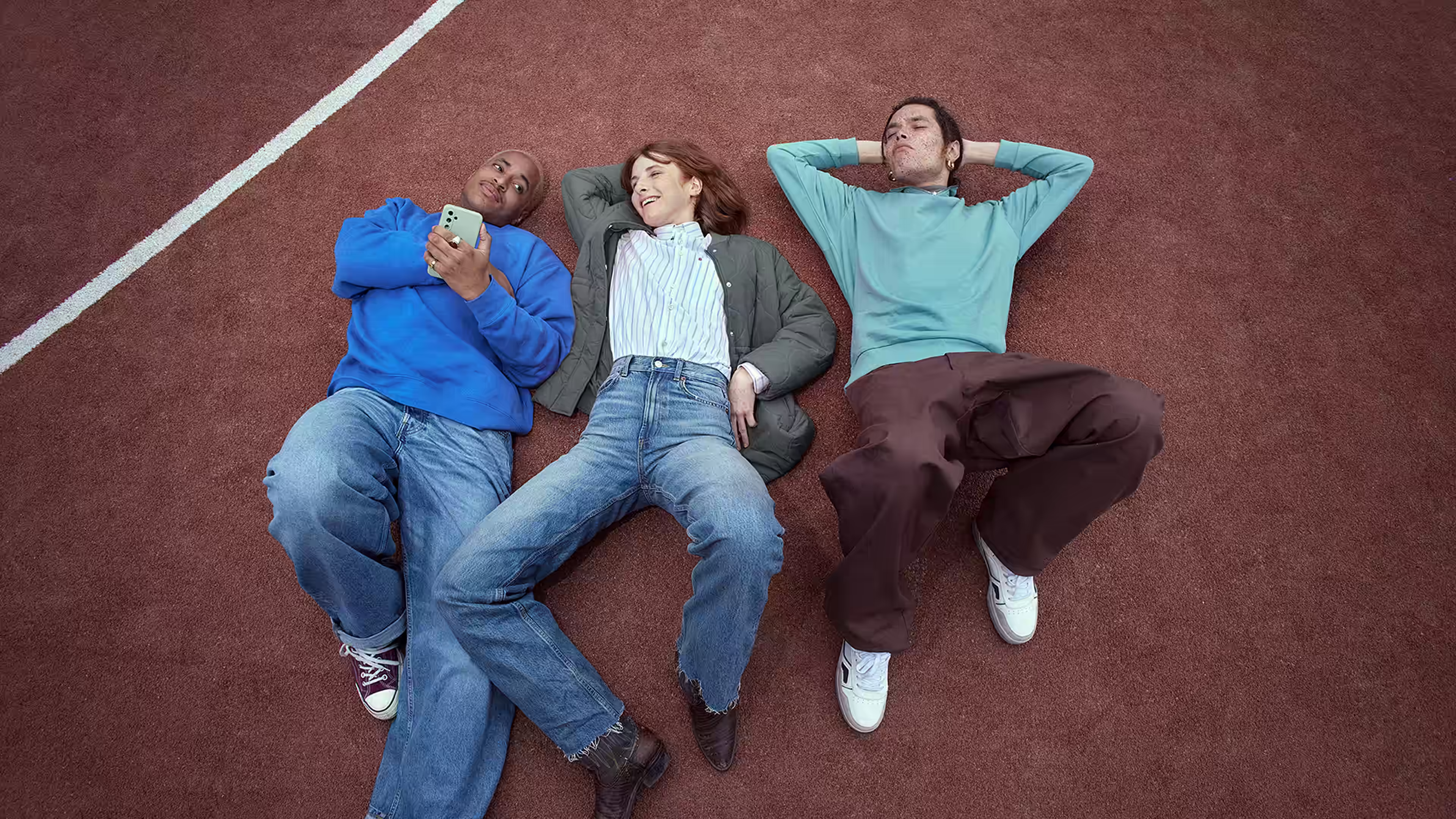 Three young adults laying on orange gravel of a basket ball court. They are laughing at a phone.