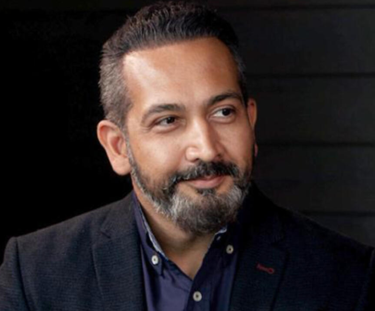 Man with short dark hair and gray beard wearing a dark blazer and navy shirt, looking to the side with a slight smile.