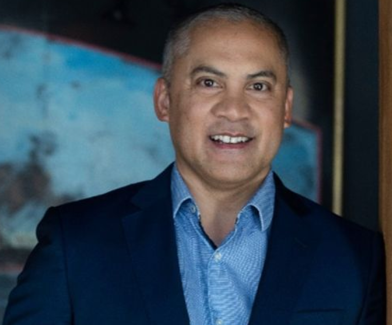 Smiling man in blue suit and light blue shirt standing indoors near a wooden beam.