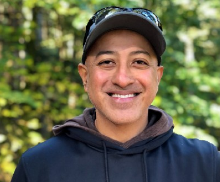 Smiling man wearing a dark hoodie, baseball cap, and sunglasses on his head outdoors with green foliage in the background.