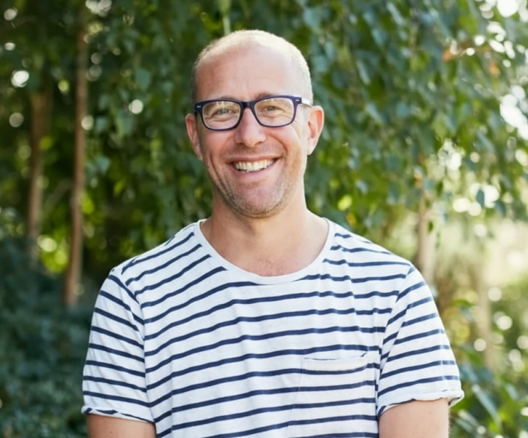 Smiling bald man with glasses wearing a white and navy striped shirt outdoors with green foliage background.
