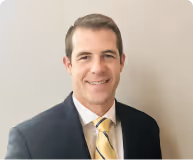 Smiling man in a dark suit with a yellow striped tie against a beige background.