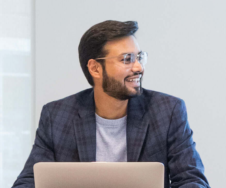 Smiling man with glasses and beard, wearing a gray shirt and checkered blazer, looking to the right.