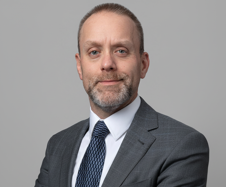 Middle-aged man with short hair and beard wearing a gray suit and blue tie, smiling slightly against a plain gray background.