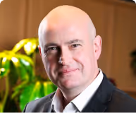 Smiling bald man wearing a black suit jacket and white shirt, with a blurred green plant and wooden wall in the background.
