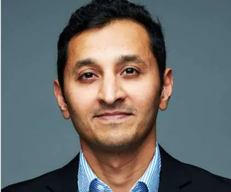 Headshot of a man with short dark hair wearing a blue shirt and dark jacket, smiling slightly against a plain gray background.