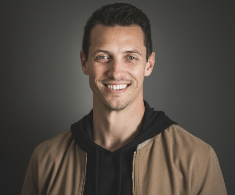 Smiling man with short dark hair wearing a brown jacket over a black hoodie against a gray background.