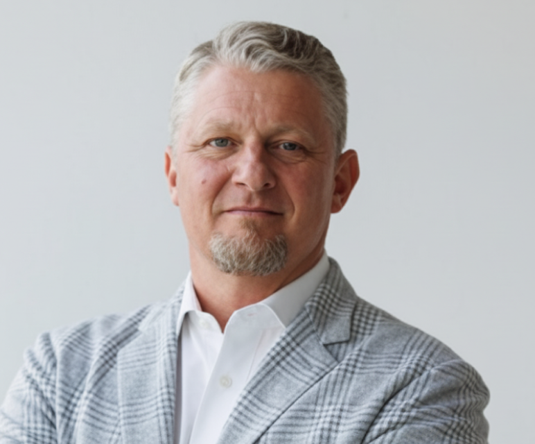 Middle-aged man with short gray hair and a goatee wearing a light gray checkered blazer and white shirt in front of a plain light background.