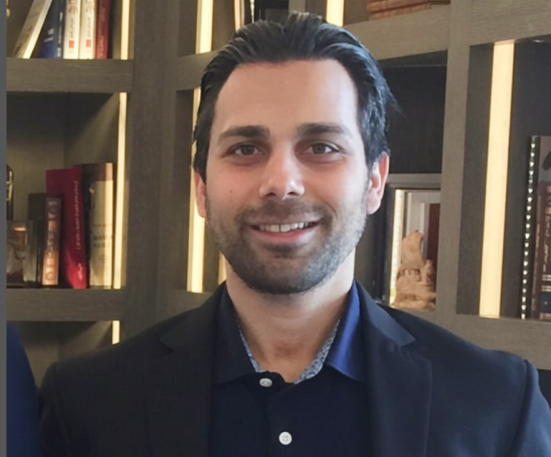 Smiling man with dark hair in a blue shirt standing in front of a bookshelf.