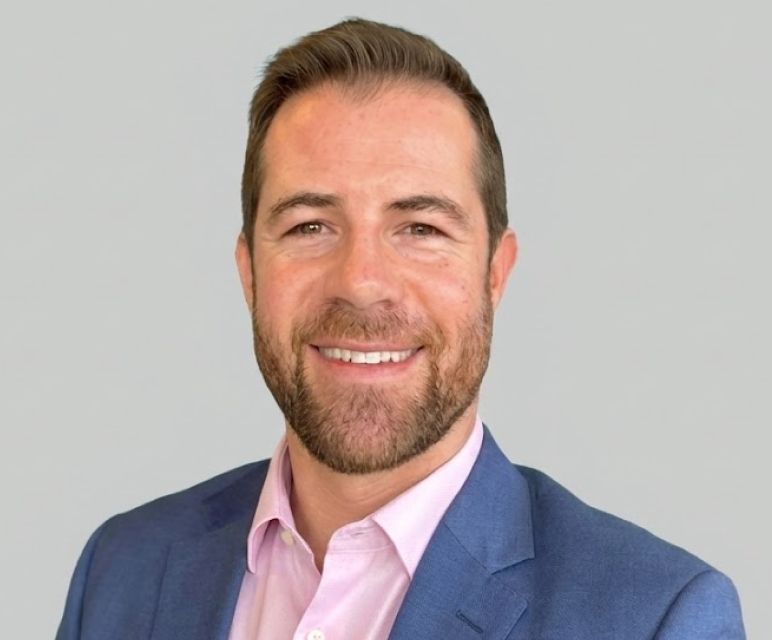 Smiling man with short brown hair, beard, wearing a blue suit jacket and light pink shirt against a light gray background.