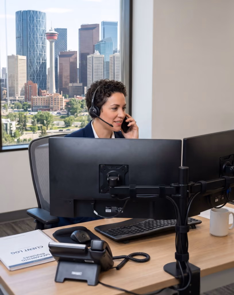 Focused customer service representative with short curly hair, wearing a headset and talking on a cell phone, while seated at a dual-monitor workstation in a modern office. On the wooden desk are a computer keyboard, mouse, a distinct desk phone, a white mug, and a spiral notebook with text that clearly reads 'CLIENT LOG' and 'Calgary and Edmonton INQUIRIES'. Through the large window behind her, a distinct city skyline featuring the Calgary Tower is visible under a clear sky. | Contact us | NewGen Cleaning Services