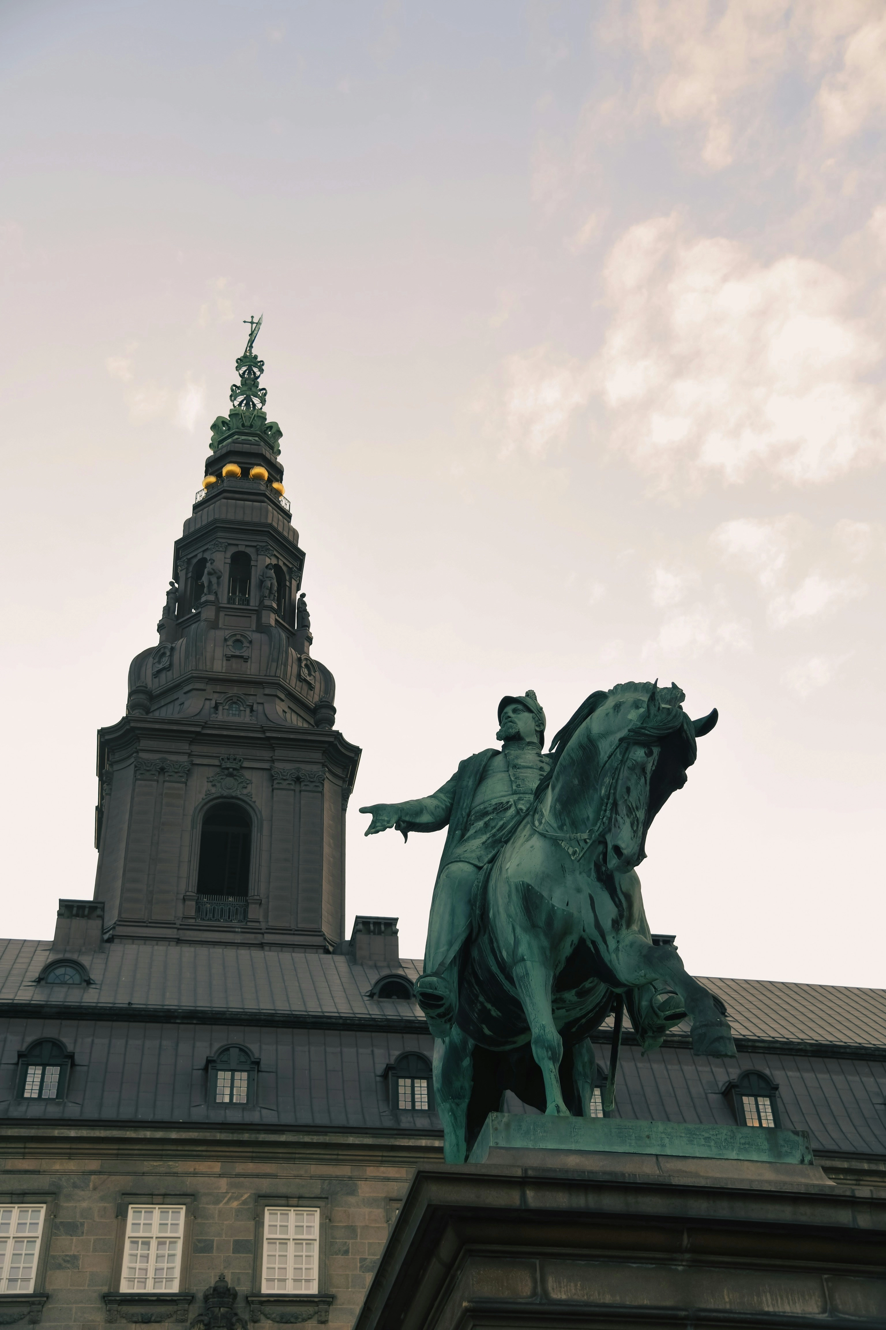 Rytterstatue foran Christiansborg Slot i København med det karakteristiske slotstårn i baggrunden under en overskyet himmel.