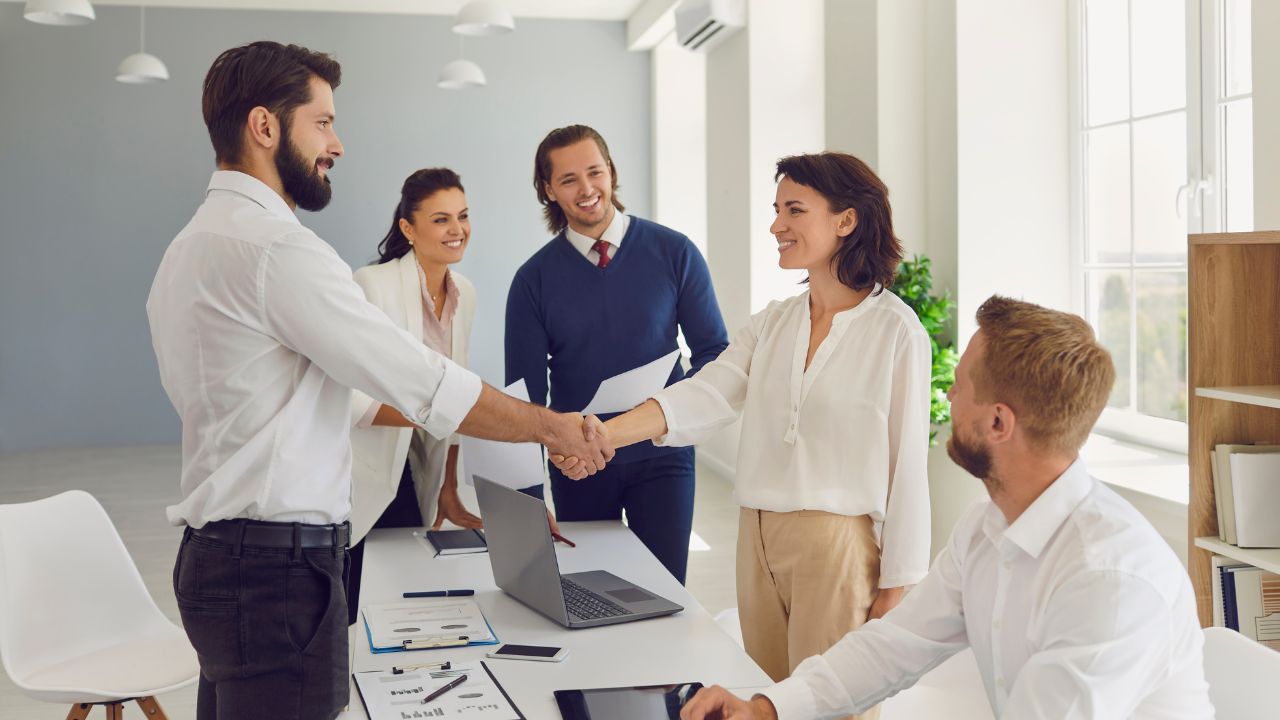 A group of people at the office. Man shaking hands with a woman, both in white shirts. Corporate envirnment.