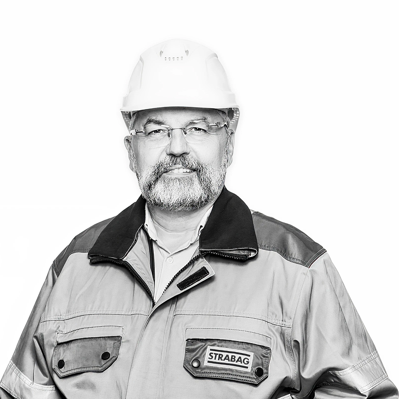 Man with beard and glasses wearing a hard hat and STRABAG work jacket posing against white.