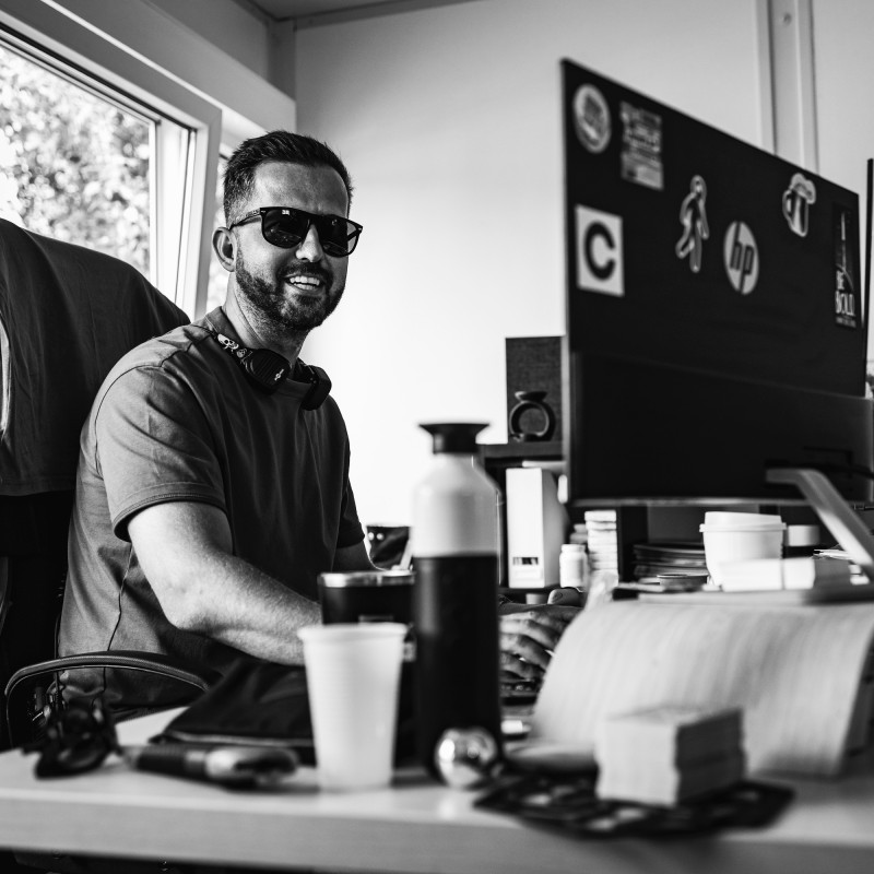 Man wearing sunglasses and headphones sitting at desk with computer, smiling in black and white photo.