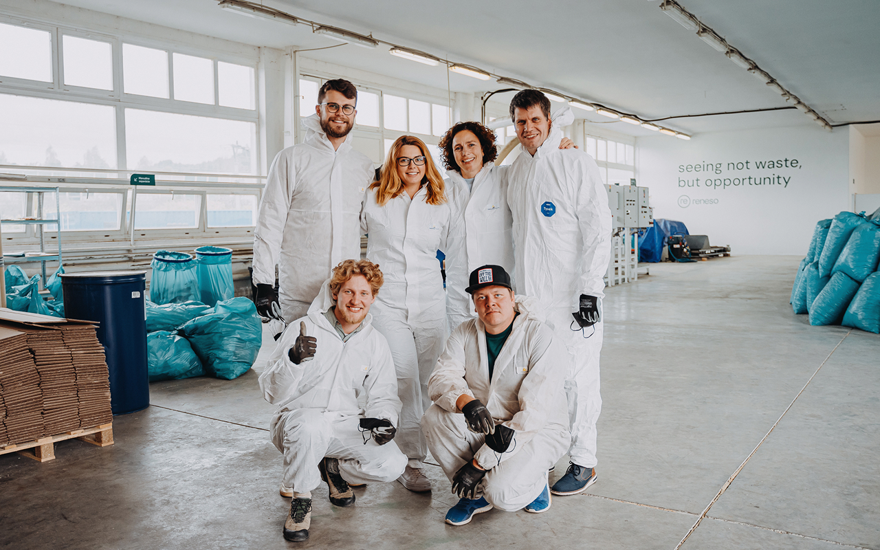 Six workers in white protective suits posing in a bright warehouse with recycling bags and cardboard.