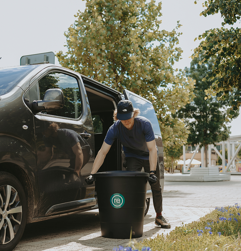 Person in gloves and cap lifting a large black bin labeled 're' next to an open van door outdoors.