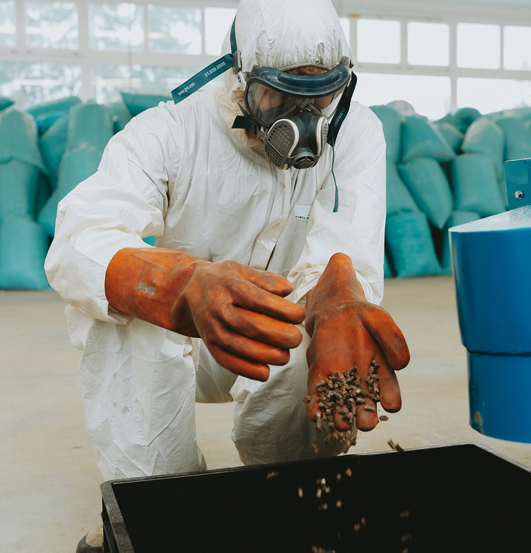 Person in protective suit, mask, and gloves handling pellets over a black container indoors.