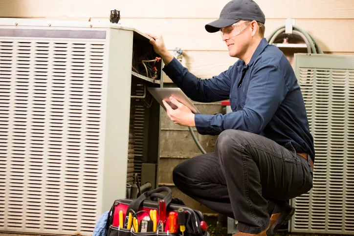 A male HVAC technician wearing a cap and safety glasses is crouching down, working on an outdoor air conditioning unit while looking at a tablet. A red toolbox is visible in the foreground.