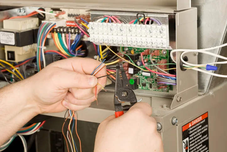 A technician's hands are shown using wire strippers to work on the complex wiring of a furnace control board.