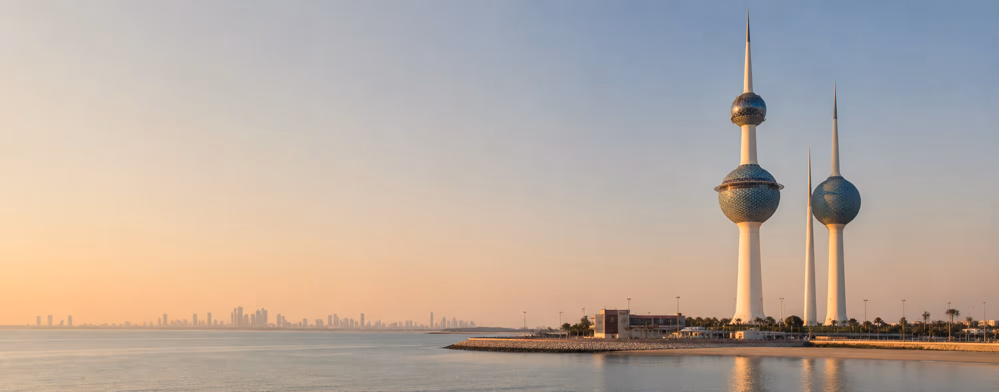 Three Kuwait Towers with sea and city skyline at sunset under a clear sky.