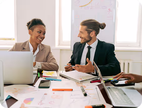 Two coworkers smiling and discussing work around a table with laptops and documents.
