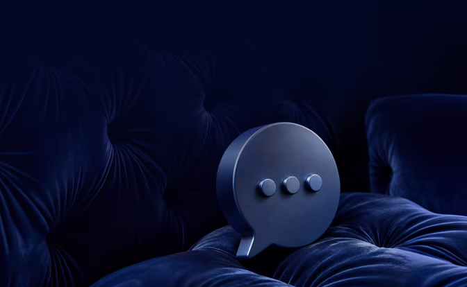 A dark metallic speech bubble icon with three raised dots in the center, placed on a deep blue tufted fabric surface.