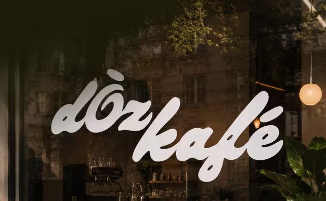 Window with white cursive text 'doz kafe' reflecting trees and buildings outside and showing interior lights and coffee equipment.