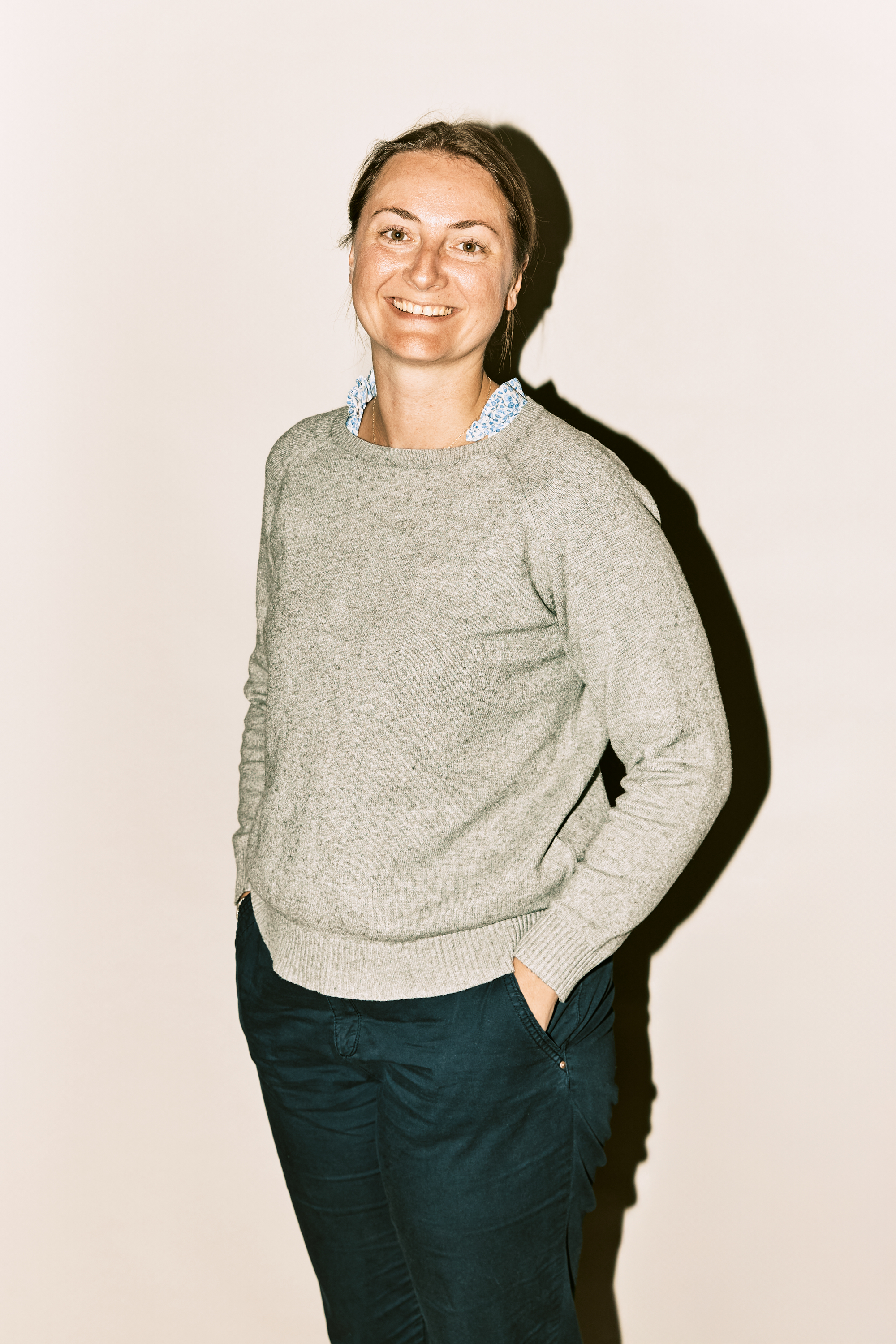 Porträt der Assistentin Renate Iten. Sie trägt ein schwarzes Oberteil und eine Brille. Sie hat schulterlange, lockige, blonde Haare und lächelt leicht mit verschränkten Armen in die Kamera.