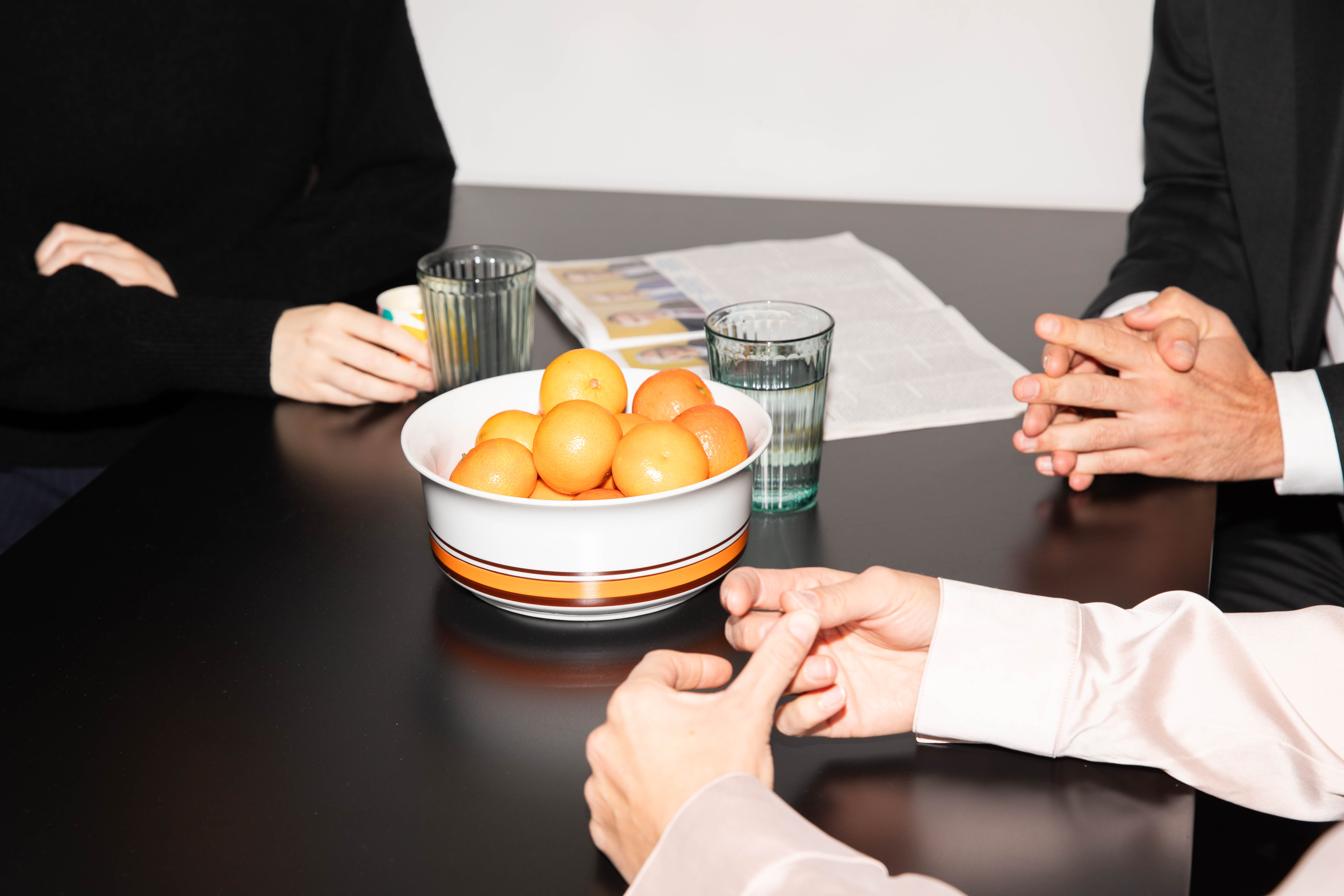 Drei Handpaare an einem dunklen Tisch, auf dem ein Schale mit Orangen steht sowie zwei Gläser und eine aufgeklappte Zeitschrift.