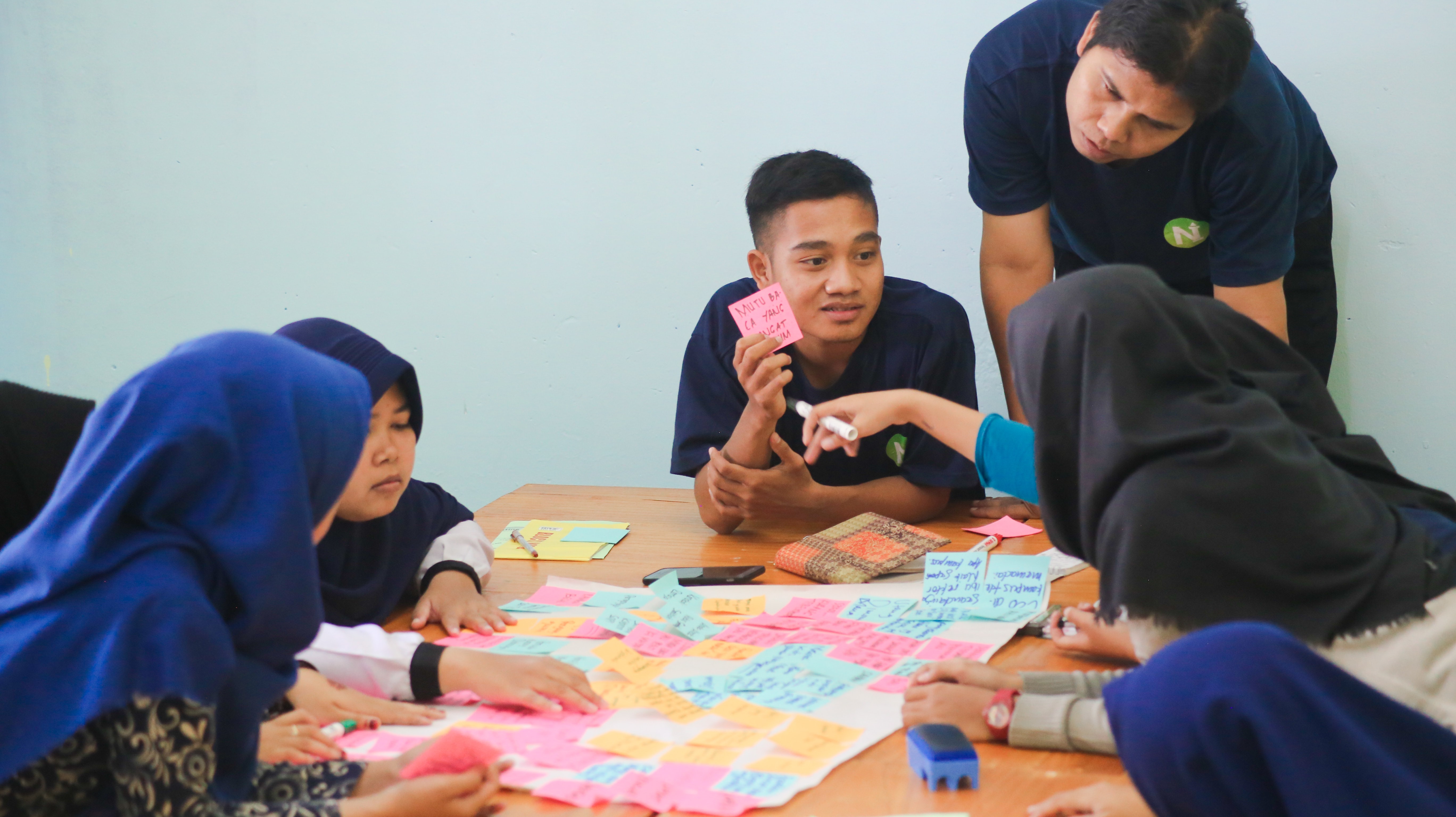 A classroom community gathers around a desk brainstorming ideas with post-it notes.