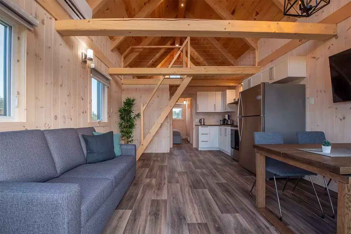 Interior of a modern wooden cabin with a gray sofa, dining table, kitchen area with stainless steel appliances, and a loft ceiling with exposed beams.