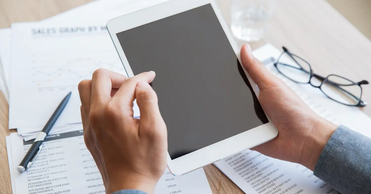 Hands holding a tablet over compliance paperwork