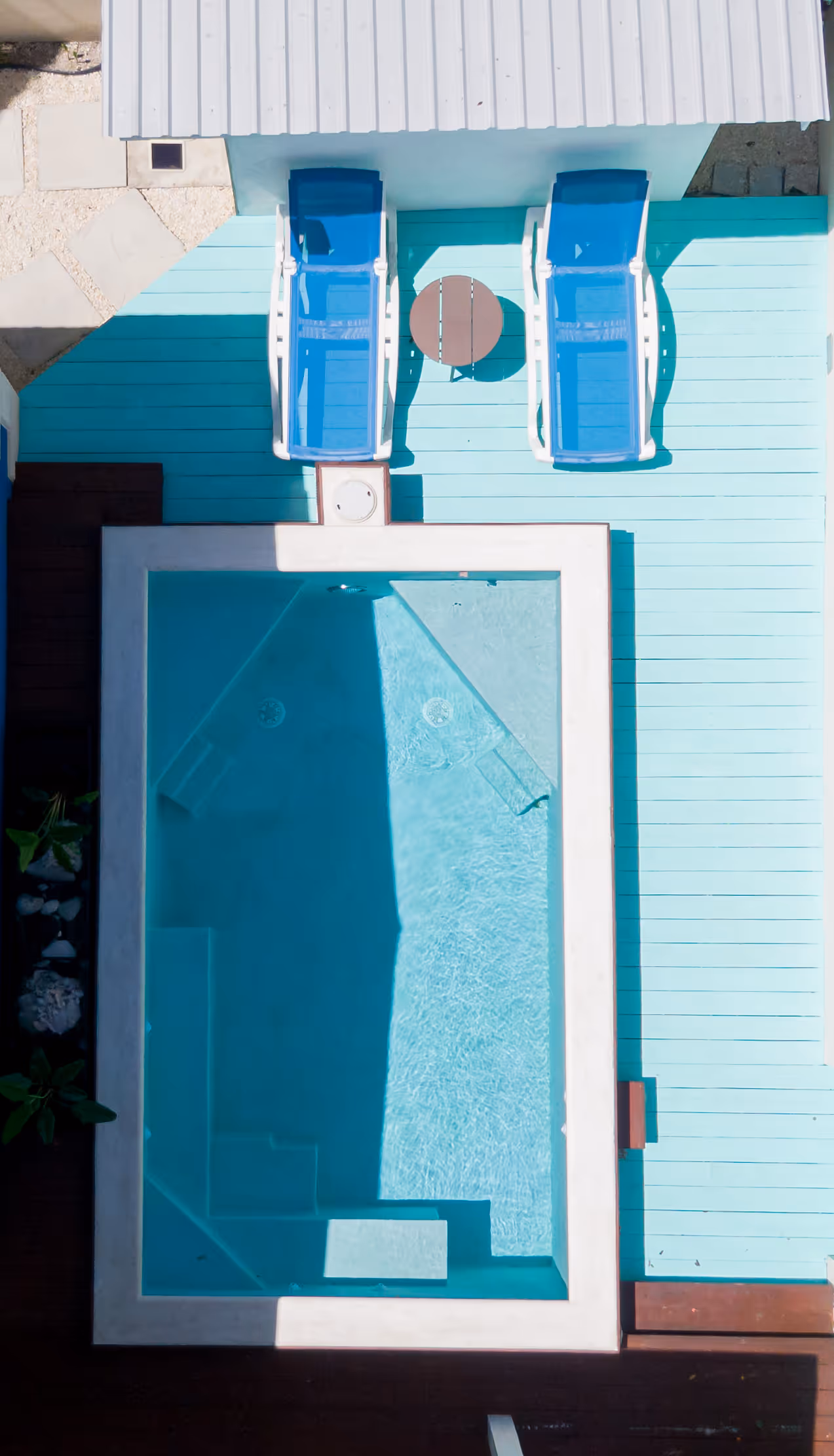 Aerial view of a small rectangular swimming pool with two blue lounge chairs and a round table on a light blue deck.