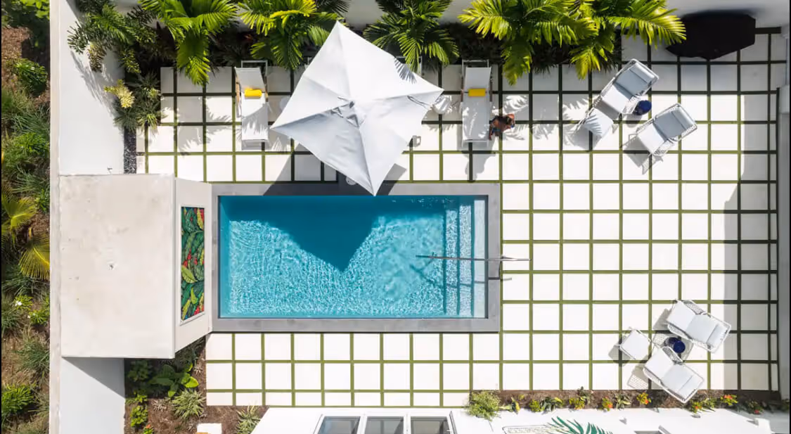 Aerial view of a rectangular swimming pool with a white umbrella, surrounded by white lounge chairs and greenery on a tiled patio.