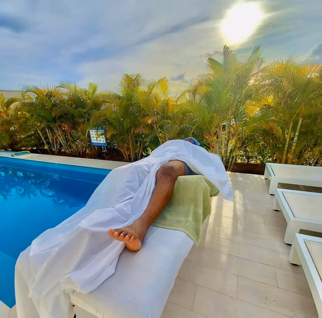 Person covered with a white sheet lying on a massage table beside a pool under a sunny sky with palm plants in the background.