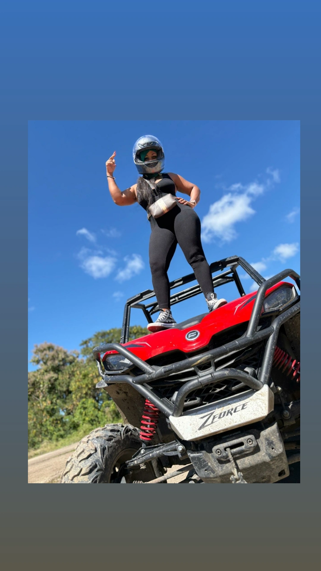 Person wearing a helmet and black outfit standing on a red ZFORCE off-road vehicle under a clear blue sky.