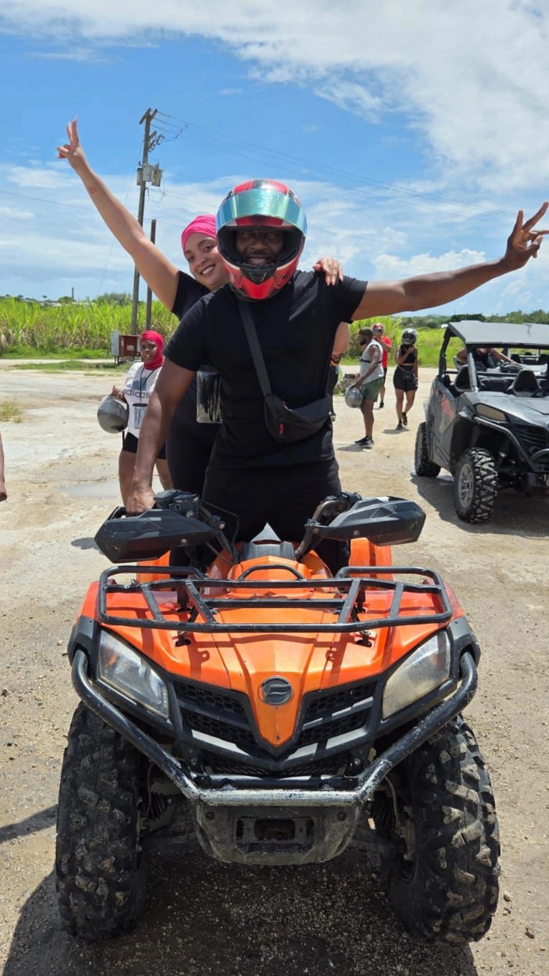 Man wearing a red helmet and black clothes sitting on an orange ATV with a woman in a pink headscarf behind him, both making peace signs.