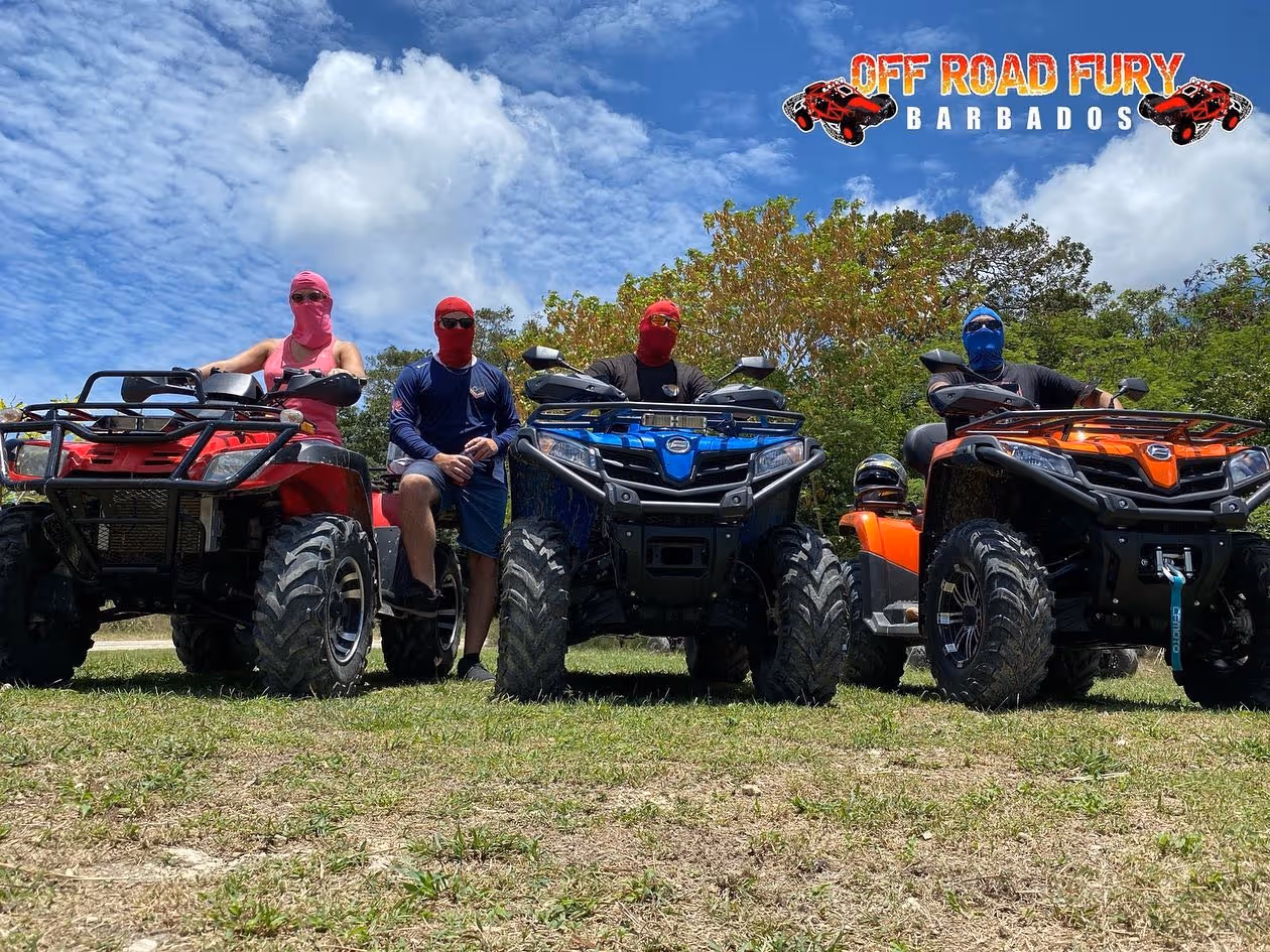Four people wearing colored face coverings sitting on red, blue, and orange off-road ATVs on grassy ground under a partly cloudy sky with 'Off Road Fury Barbados' text and logo above.