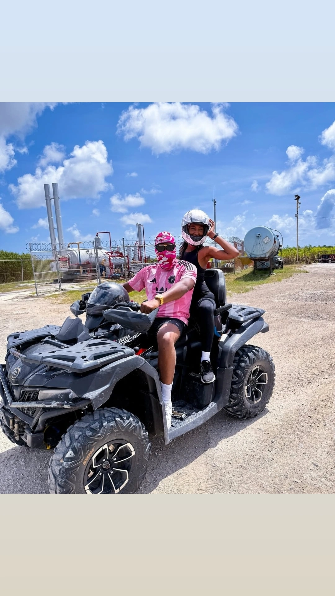 Man and woman wearing helmets sitting on a black all-terrain vehicle on a dirt road with industrial tanks and fences in the background under a partly cloudy sky.