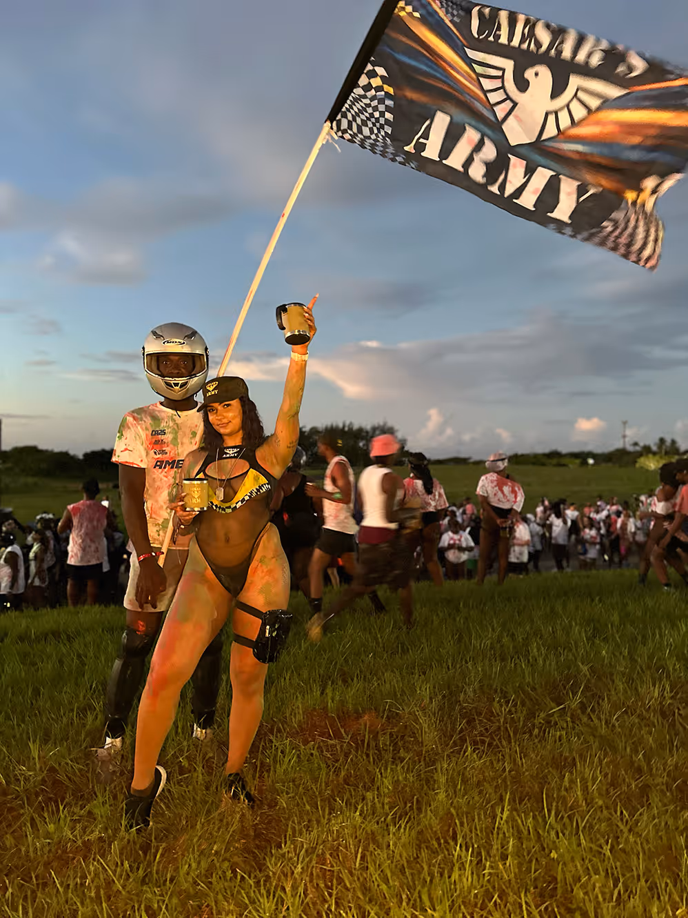 Woman in a black mesh outfit holding a drink and a Caesar's Army flag stands beside a helmeted man on a grassy field during an outdoor event with a crowd in the background.