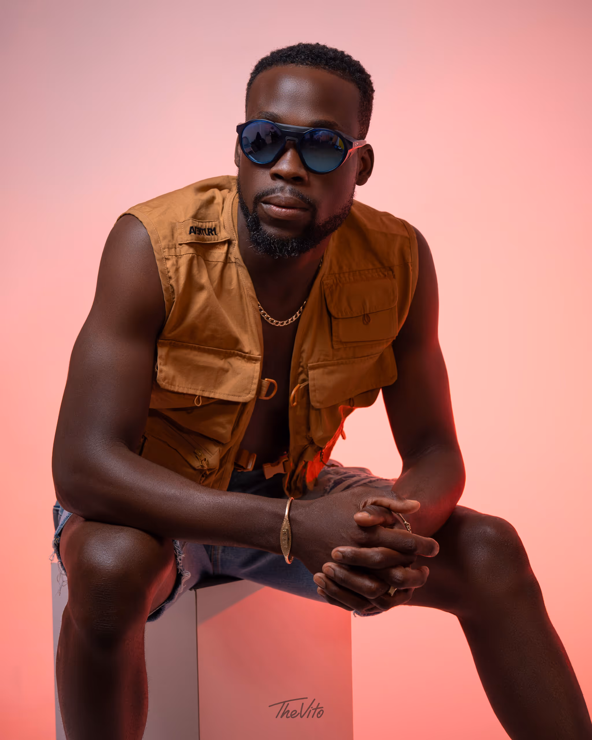 Man wearing sunglasses, a brown sleeveless vest, and denim shorts sitting on a white cube against a pink background.