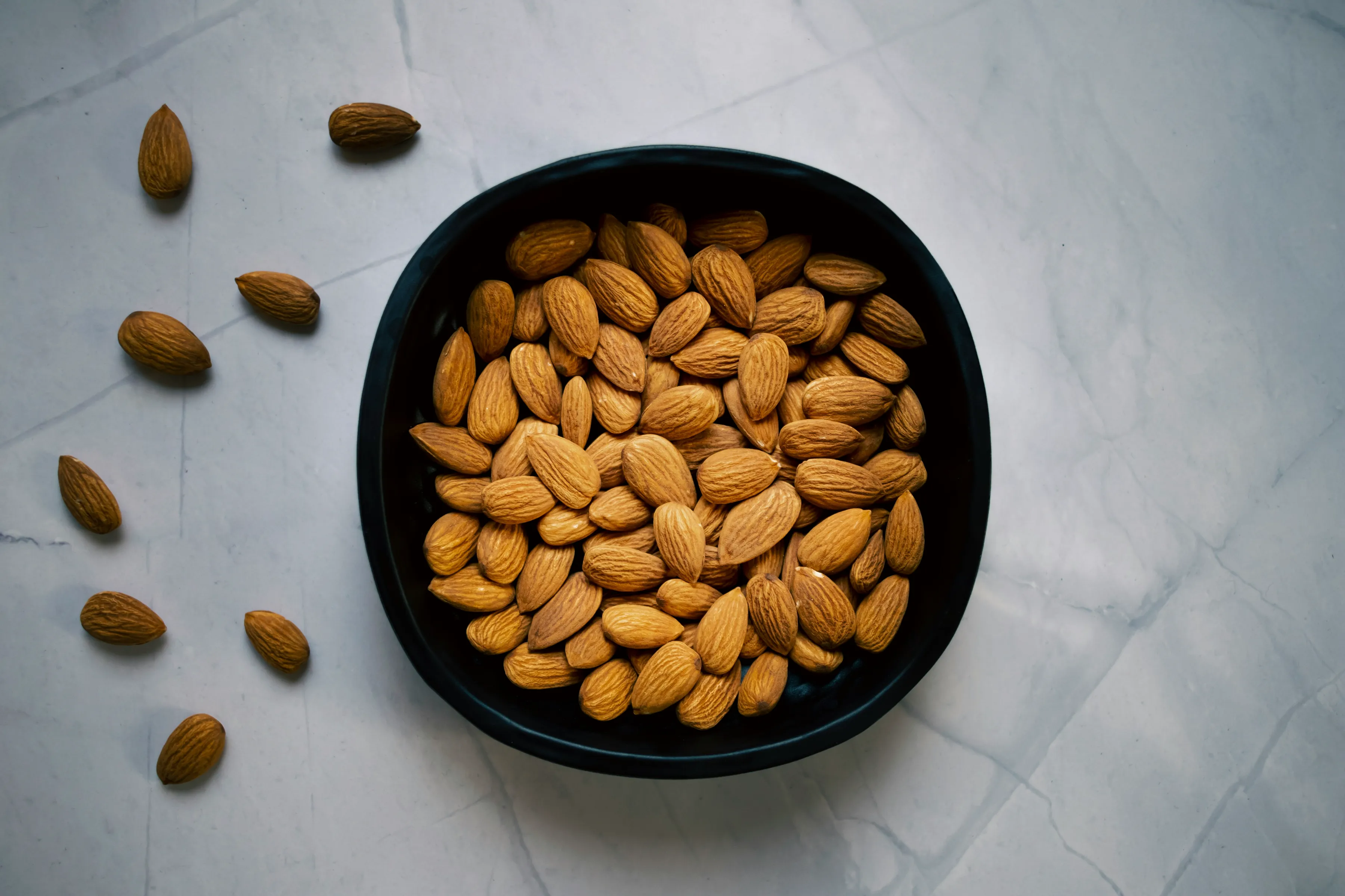 A matte black bowl filled with raw California almonds sits on a light marble surface at Sharon Naraghi Farms. A few nuts are scattered around the bowl, emphasizing freshness and natural texture—part of the farm’s premium, sustainably grown San Joaquin Valley crops.