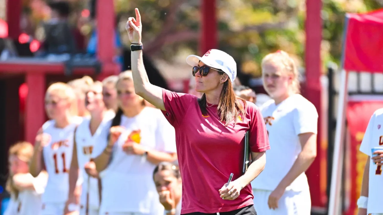 Female lacrosse coach in a maroon shirt and white hat signaling a play with a raised finger while holding a clipboard on the sideline.
