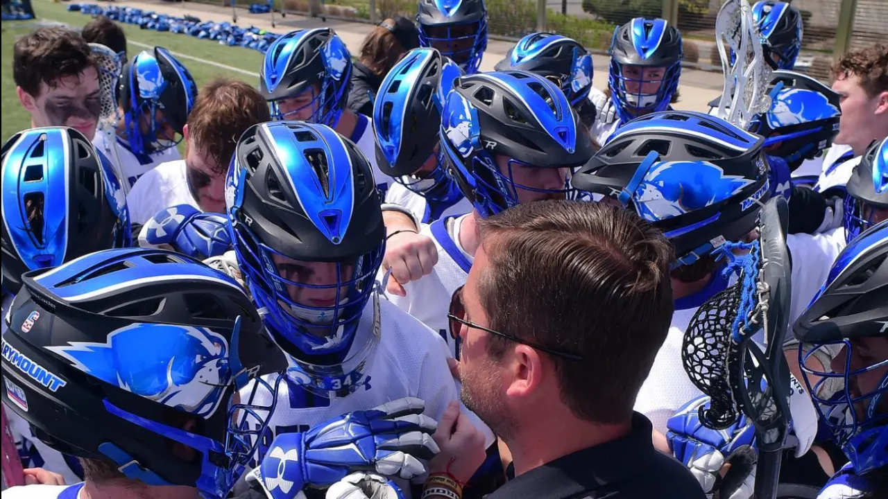 Marymount men's lacrosse coach addressing players in blue chrome helmets during a focused team huddle on the field.